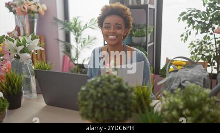 An african american woman entrepreneur managing her flower shop business with a computer and documents, surrounded by lush plants. Stock Photo