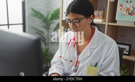 A young, attractive, brunette, hispanic female doctor works diligently in a clinic room while wearing a white coat and stethoscope, examining medical Stock Photo