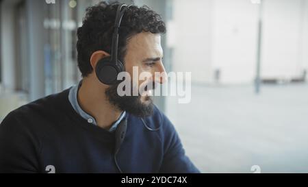 A bearded man wearing headphones in a modern office setting, looking away thoughtfully. Stock Photo