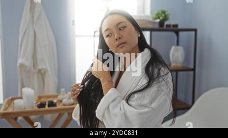 Beautiful young chinese woman in a spa room, wearing a white robe, combing her long hair, surrounded by a serene and tranquil beauty center interior. Stock Photo