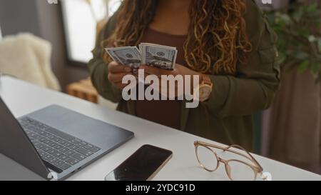 Woman counting money in a home interior decor setting with a laptop, phone, and glasses, suggesting a blend of work and personal life Stock Photo
