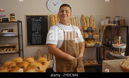 Young hispanic man in a bakery shop posing confidently indoors with bread and pastries displayed around him Stock Photo