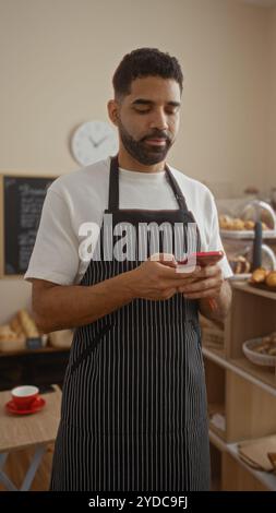 Young hispanic man in a bakery checking his phone while wearing an apron Stock Photo