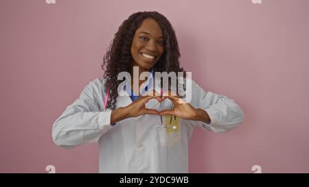 A beautiful young african american woman with curly hair smiles while making a heart shape with her hands, standing against a pink background in a med Stock Photo
