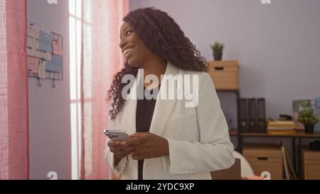 A beautiful young african american woman with curly hair smiles in a police detective's office holding her phone, standing beside a window with pink c Stock Photo