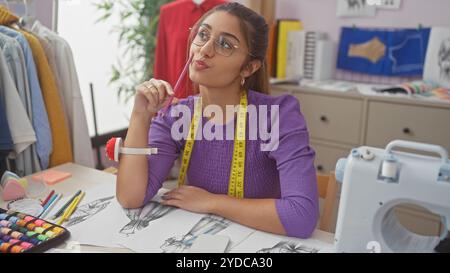 A thoughtful young woman with glasses sits in a colorful atelier, surrounded by fashion sketches, a sewing machine, and fabric. Stock Photo