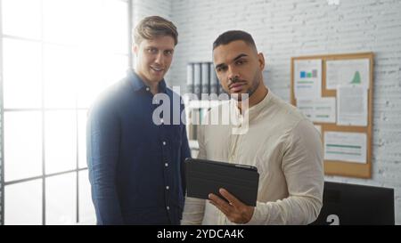 Men standing in an office holding a tablet, showcasing a business partnership between two handsome male employees in a workplace setting. Stock Photo