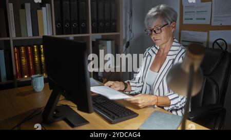 Woman working in an office, middle-aged, short grey hair, holding documents, sitting at a desk with a computer, surrounded by shelves filled with file Stock Photo