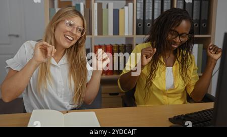Young women smiling and dancing joyfully in an office while working on a project together, surrounded by books and files Stock Photo