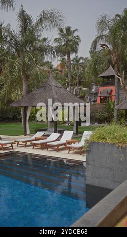 Luxurious tropical resort pool with loungers and thatched cabanas surrounded by palm trees Stock Photo