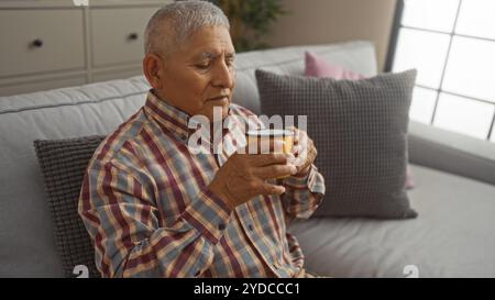 A mature hispanic man with grey hair, wearing a plaid shirt, sitting in a cozy living room, holding a coffee cup with both hands, enjoying a serene mo Stock Photo