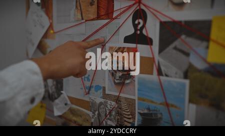 A young asian man points at a clue on a detective's evidence board in a dimly-lit police station. Stock Photo