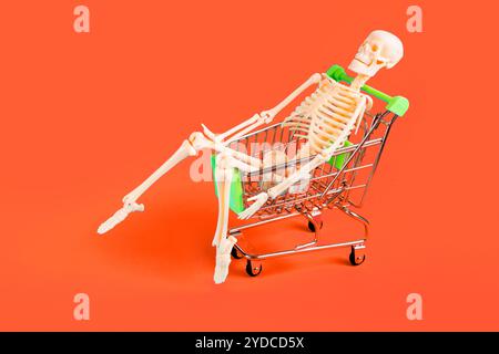 Playful skeleton sitting in a shopping cart against a vibrant orange background. Halloween goods promotion concept. Stock Photo