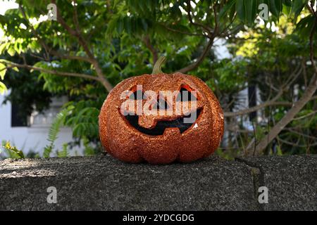 Orange sparkle covered jack o' lantern sitting atop a stone wall, in front of a suburban home's yard, during the day Stock Photo