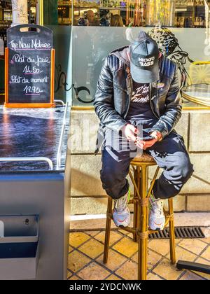 Young man sitting on high stool on café terrace head bent down using smartphone - Tours, Indre-et-Loire (37), France. Stock Photo