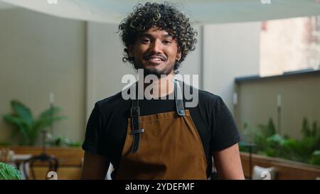 Happy cafe worker Latino American ethnic man in apron guy waiter male cleaner barista working outside cafeteria sweeping leaves broom cleaning looking Stock Photo