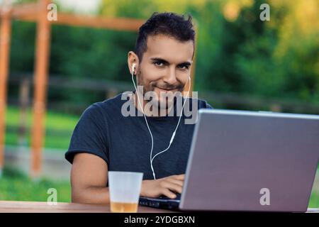 Young freelancer smiling and working remotely on his laptop in a park, wearing earphones and enjoying a refreshing drink Stock Photo