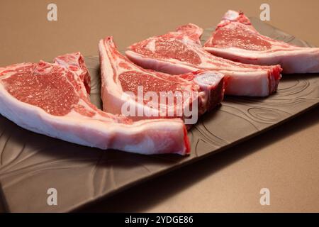 A close-up of four raw lamb chops on a black plate shows their marbled pink meat and fat layers The cuts are fresh and neatly arranged ready for grill Stock Photo