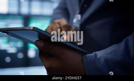 Software developer doing yearly maintenance using tablet to prolong data center equipment life span. Extreme close up of worker in server hub monitoring server infrastructure using device, camera B Stock Photo