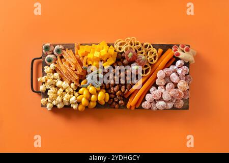 Halloween sweet charcuterie board with pumpkin, popcorn, dried fruits, color chocolate candy, spider, crackers on orange background. Festive food for Stock Photo