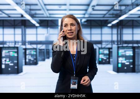 IT manager overseeing operations in cutting edge data center, coordinating tasks during phone call. Woman in server room managing systems and communicating via smartphone Stock Photo