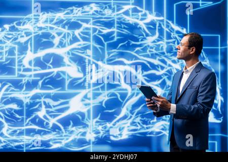 Data scientist analyzes AI brain models on tablet with large digital brain displayed behind. IT expert using machine learning to better understand and develop artificial intelligence Stock Photo