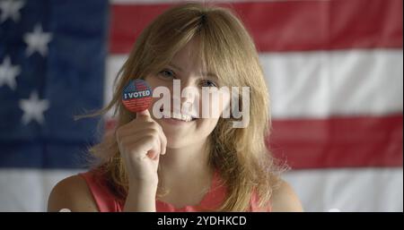 Close up, red-haired young woman holds up round 'I voted' sticker on thumb near face. Shallow depth of field, US flag behind Stock Photo