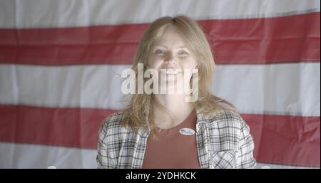 Close up, red-haired young woman wearing "I voted" sticker and smiling. Large United States flag on back wall in background Stock Photo
