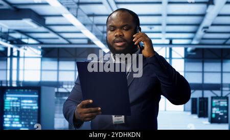 IT professional looking over data center checkup checklist paperwork, talking on phone in server farm. African american man surrounded by server rigs, chatting during telephone call, camera A Stock Photo
