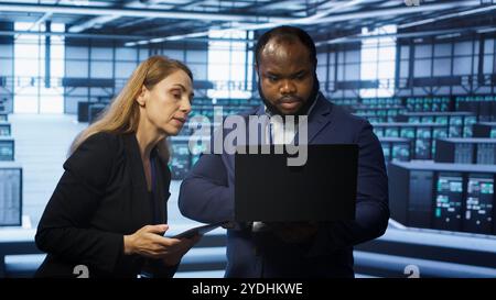 System administrators in data center overseeing server operations using notebook and tablet. Colleagues brainstorming in server hub, ensuring optimal performance and data integrity, camera A Stock Photo