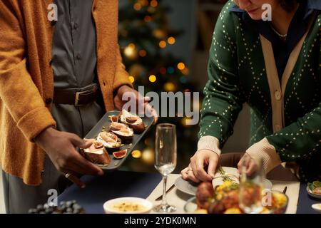 Cropped shot of man serving plate of tartines with figs and ricotta on dining table and woman decorating dishware for festive Christmas dinner Stock Photo