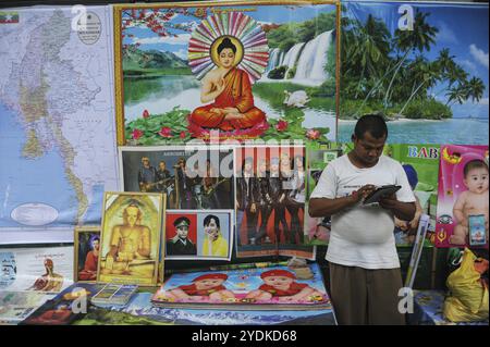 13.08.2014, Yangon, Myanmar, Asia, A street vendor stands in front of his stall with posters at a street market in the city centre and looks at his iP Stock Photo