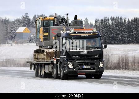 Black Volvo FMX truck Lonnqvist Oy hauls Volvo excavator on flatbed trailer along highway 52 on a day of winter. Salo, Finland. December 27, 2021 Stock Photo