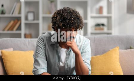 Bored millennial generation man sitting on sofa, feeling lonely at home. Unhappy young male suffering from negative thoughts, having depressive mood Stock Photo