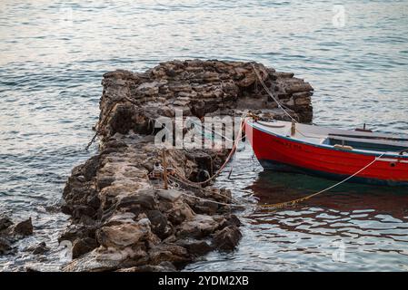 A red wooden boat moored tied with ropes to rocky reefs and skerries at Zakynthos in Greece in the early morning. Sea is calm, the scene is peaceful. Stock Photo