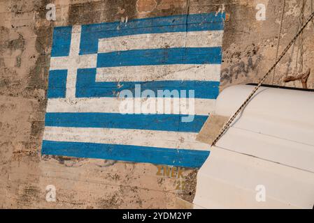 Greek flag painted blue and white on a cement wall close to a boat hauled out of water and tied with ropes. Stock Photo