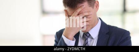 Depressive man in formal suit covers face with palm in office Stock Photo