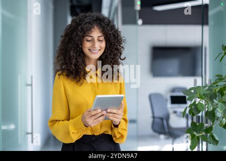 Latin American businesswoman in office using tablet, representing professionalism. Smiling in yellow blouse, standing in hallway with digital device, symbolizing technology in workplace. Stock Photo