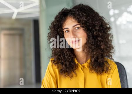 Latin American businesswoman with curly hair wearing yellow blouse. Expresses professionalism, confidence, approachability in modern office. Ideal for corporate, leadership, business themes. Stock Photo