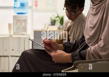 Muslim woman writing notes on tablet in modern office while colleague focuses on screen behind. Desk and office supplies are visible in bright workspace Stock Photo