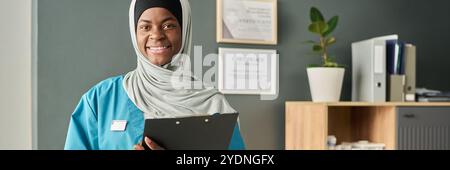 Muslim medical professional in healthcare uniform holding clipboard, smiling warmly. Documents in background indicating a healthcare setting with certificates and folders Stock Photo