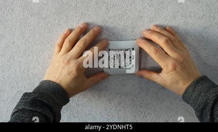 worker fixing or maintaining an electrical outlet or light switch attaching a white plastic frame with two hands against gray wallpaper Stock Photo