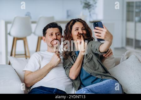 Young couple is having fun taking a selfie on the sofa at home, making mustaches with their hair and enjoying a relaxing day together Stock Photo