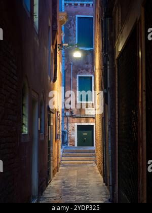 Tight alleyway in Venice illuminated by a warm street lamp as the wet cobbled floor reflects the cityscape above between the buildings. Stock Photo