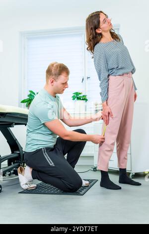 Young woman with lower back pain in physical therapy. Chiropractor examining female patient using measuring tape to determine mobility level. Stock Photo