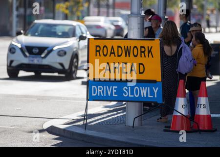 A 'Roadwork Ahead' sign with 'Drive Slowly' instruction on a busy urban street, with pedestrians and a car in the background. Stock Photo