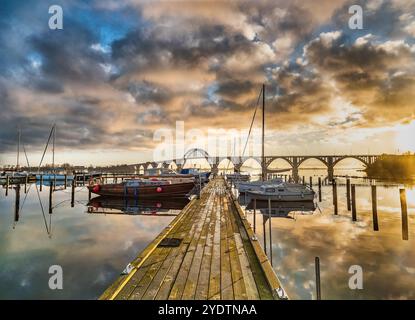 Dramatic sunset sky over a calm harbor, with boats docked along the pier and a scenic Queen Louise bridge in the background. A tranquil waterfront set Stock Photo