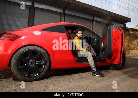 A confident man in a mustard yellow shirt sits casually inside a sleek red Audi TT with black rims, parked against a rugged industrial backdrop. The open car door and his intense gaze add to the bold, urban vibe of the scene, blending luxury with an edgy, street-style aesthetic. Stock Photo