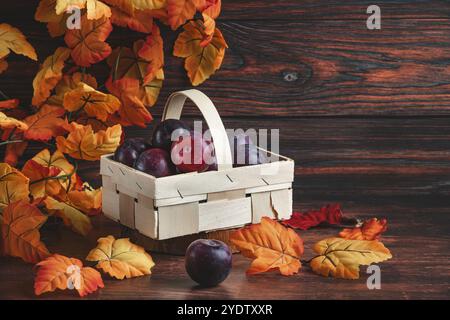 Plums in a wooden basket surrounded by orange autumn leaves Stock Photo
