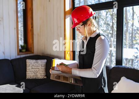Caucasian woman inspector during pre-purchase inspection, she wearing a red hardhat and protective eyewear and using a clipboard, home interior review Stock Photo